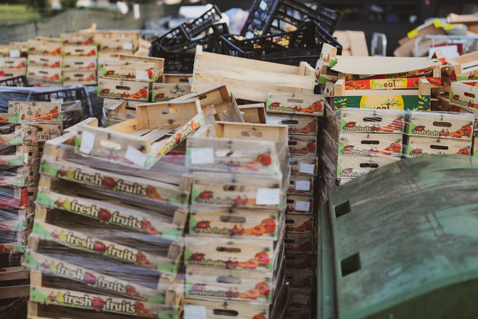 A large outdoor scene featuring numerous cardboard boxes labeled 'fresh fruits' stacked on top of each other, with some boxes appearing empty or partially collapsed. The boxes are decorated with colorful fruit illustrations and are placed on a surface that may be a pavement or industrial area. In the background, there are black plastic crates and a green, weathered, curved object that resembles a large storage container or recycling bin lid. The environment suggests a setting where waste or recyclable materials are temporarily stored before collection, emphasizing the importance of proper rubbish disposal. The lighting appears natural, highlighting the worn textures of the cardboard and the variety of stacking arrangements, which reflects an active waste management or rubbish removal process possibly handled by an independent service such as Rubbish Removal Tooting.