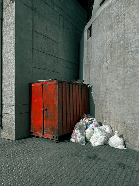 A red metal skip with a weathered finish and vertical ridges is placed against a dark green wall and a light grey concrete wall at a street corner. The skip's surface shows signs of rust and paint chipping. Adjacent to it, several white plastic rubbish bags filled with mixed waste are piled on the dark brick pavement, some leaning against the skip. In the background, the dark green wall extends upward, and part of a grey building facade with a small rectangular window is visible. The environment appears to be an urban outdoor area, possibly an alley or side street, where private waste collection or rubbish removal services, such as those offered by Rubbish Removal Tooting, might be engaged to handle disposal outside regular council collection. The lighting is natural, suggesting daytime, with no significant shadows or reflections. The scene depicts typical arrangements for local waste management and alternative rubbish handling at a commercial or residential property boundary.