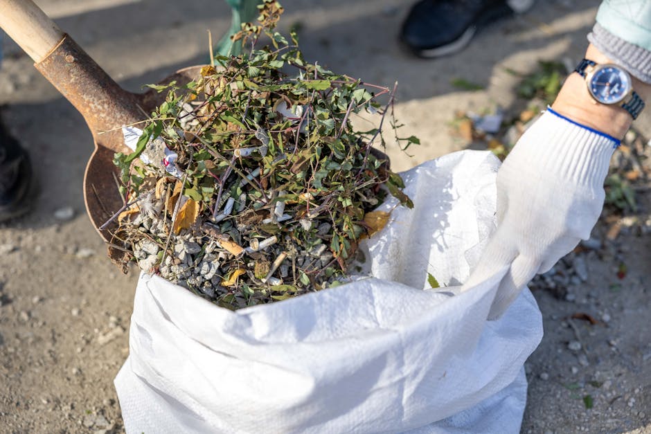 A person wearing a white glove is holding open a large white rubbish sack filled with garden debris, including small branches, leaves, and gravel. The debris includes green leafy stems and some dried brown leaves, with the gravel appearing grey and rough-textured. The person is standing outdoors on a paved or gravel surface, with part of a dark shoe visible in the background. A manual shovel with a rusted metal blade and wooden handle is positioned nearby, partly obscured. This scene depicts an on-site waste collection or garden rubbish removal process, consistent with private disposal services offered by Rubbish Removal Tooting, emphasizing efficient and professional clearance of garden waste at the property.