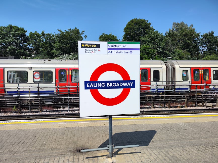 A close-up view of a station sign at Ealing Broadway indicating train services and directions, positioned centrally on a metal pole. Behind the sign, there is a modern train carriage with a silver exterior, featuring a red and blue livery along its windows and doors, which are closed. The train is stationary on the railway tracks, framed by a black metal fence with horizontal bars running parallel to the platform edge. The platform surface is made up of interlocking concrete pavers, and the background shows a line of lush green trees under a bright blue sky with no visible cloud cover. The scene suggests an active urban railway station environment, with clear natural lighting highlighting the details of the sign, train, and surrounding environment, aligning with themes of transport infrastructure and public travel, similar to elements encountered in local transport hubs like those associated with rubbish removal and waste disposal services near busy stations.