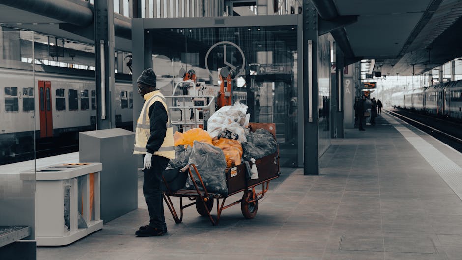 A person wearing a white glove is holding open a large white rubbish sack filled with garden debris, including small branches, leaves, and gravel. The debris includes green leafy stems and some dried brown leaves, with the gravel appearing grey and rough-textured. The person is standing outdoors on a paved or gravel surface, with part of a dark shoe visible in the background. A manual shovel with a rusted metal blade and wooden handle is positioned nearby, partly obscured. This scene depicts an on-site waste collection or garden rubbish removal process, consistent with private disposal services offered by Rubbish Removal Tooting, emphasizing efficient and professional clearance of garden waste at the property.